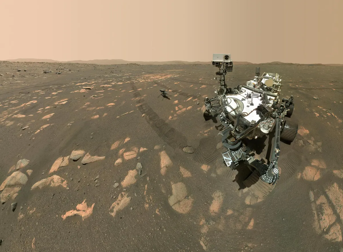 A rover's selfie on a dusty plain, with a small helicopter standing behind it